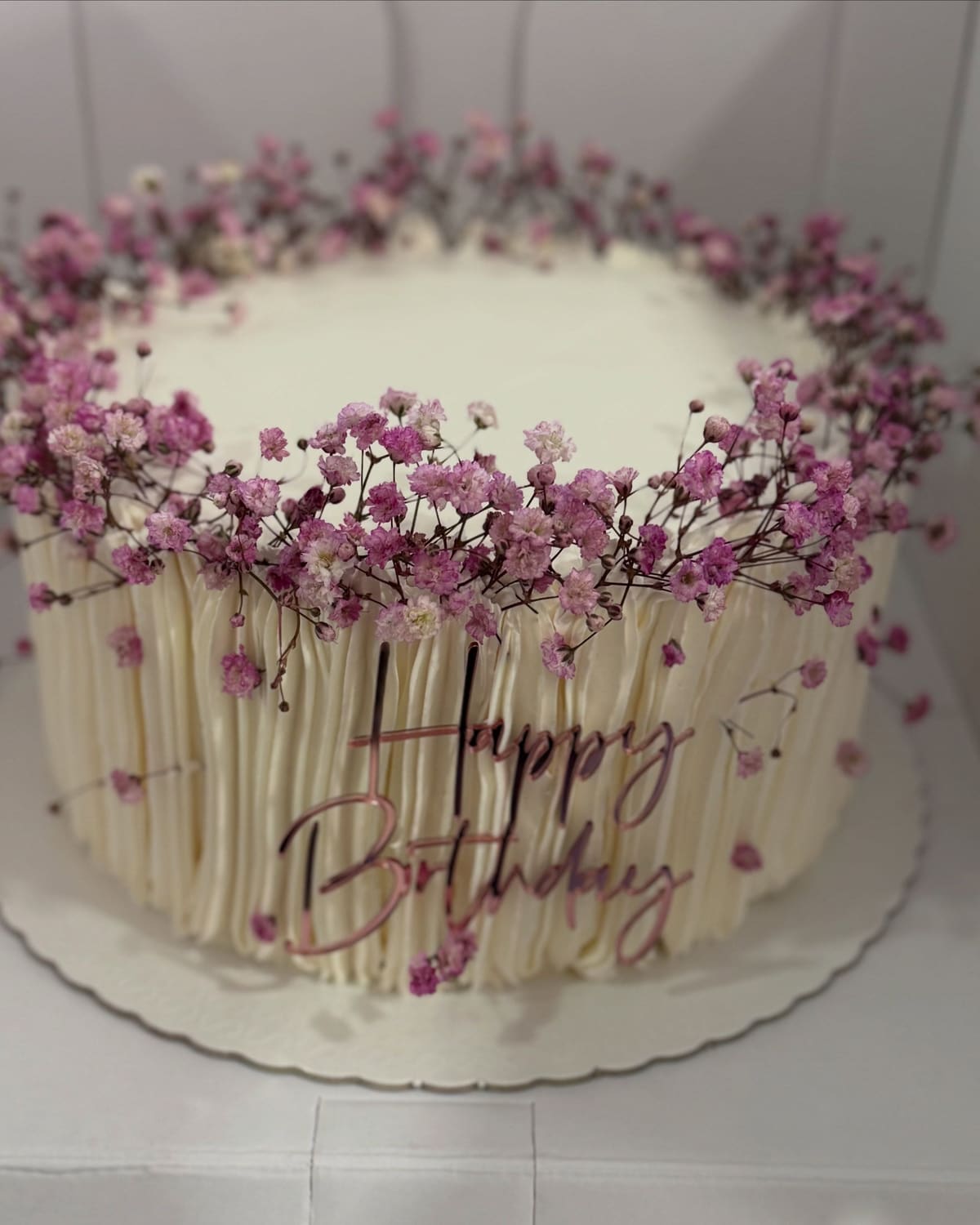 Bolo de aniversário branco com laterais texturizadas, flores lilás no topo e “Happy Birthday” escrito na lateral
